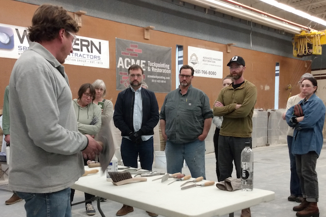 A person behind table describing the brick mortar tools on the table to several people