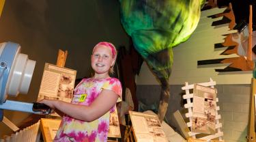 A girl holding a directional fan standing in front of a cloth tornado and damaged house.